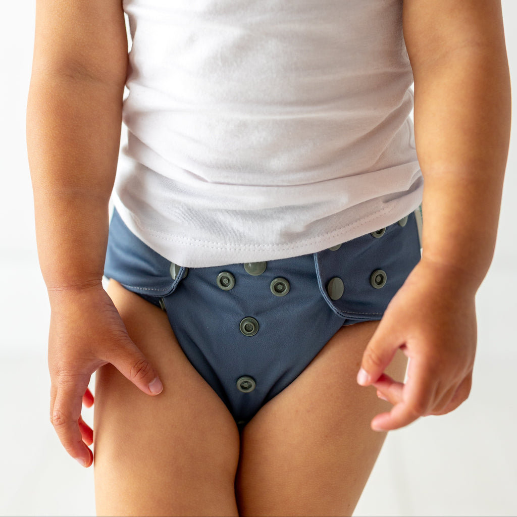 A child wearing a white tank top and blue reusable cloth diaper with green buttons, shown against a plain light background.