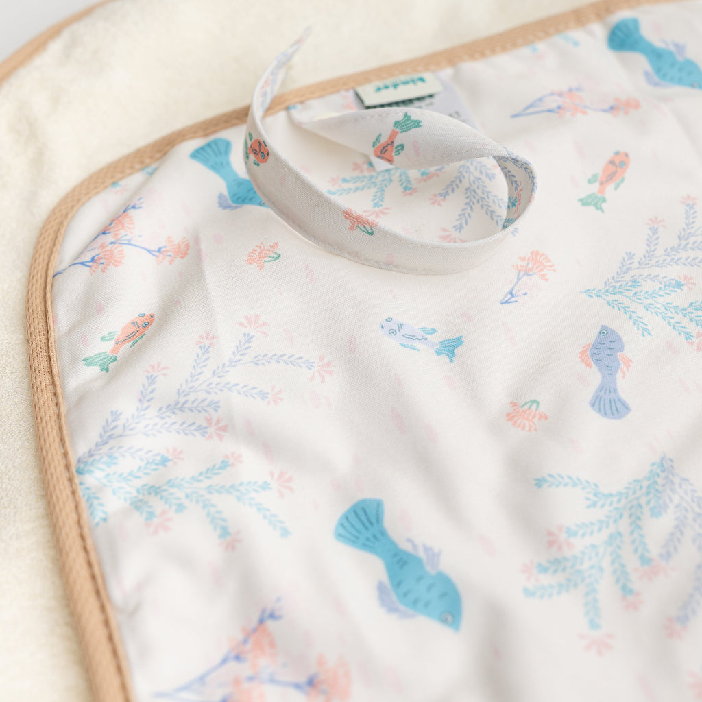 A close-up of a baby blanket featuring a white background with a pastel pattern of blue fish, pink sea anemones, and light blue seaweed. The blanket has a beige stitched border and a white fabric loop with a small green and blue brand tag.